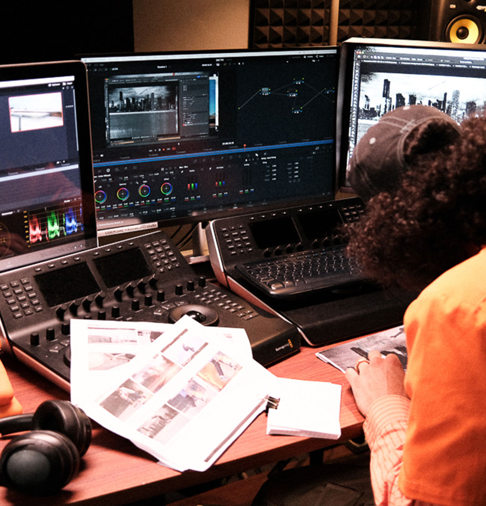 Three monitors are in front on desk. Two mixing consoles in front of person looking down at paper. Paper on table