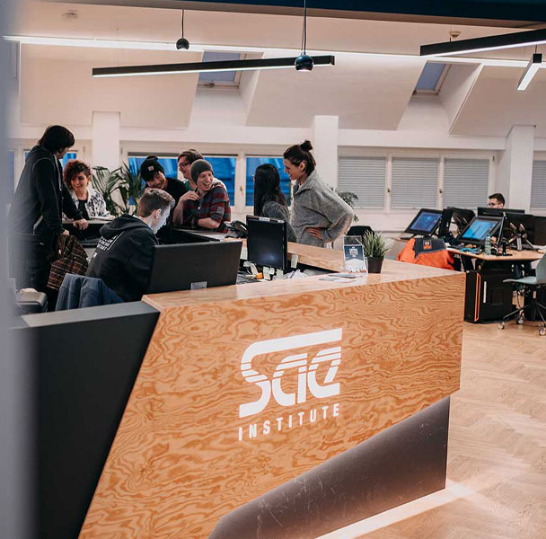 Office setting with many people around a front desk with SAE Institute logo on front of desk. Desks and monitors set up in the background.