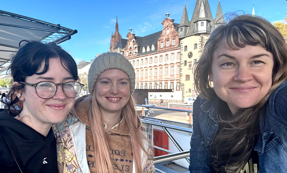 Three women in a selfie taken on a boat with historic buildings in background