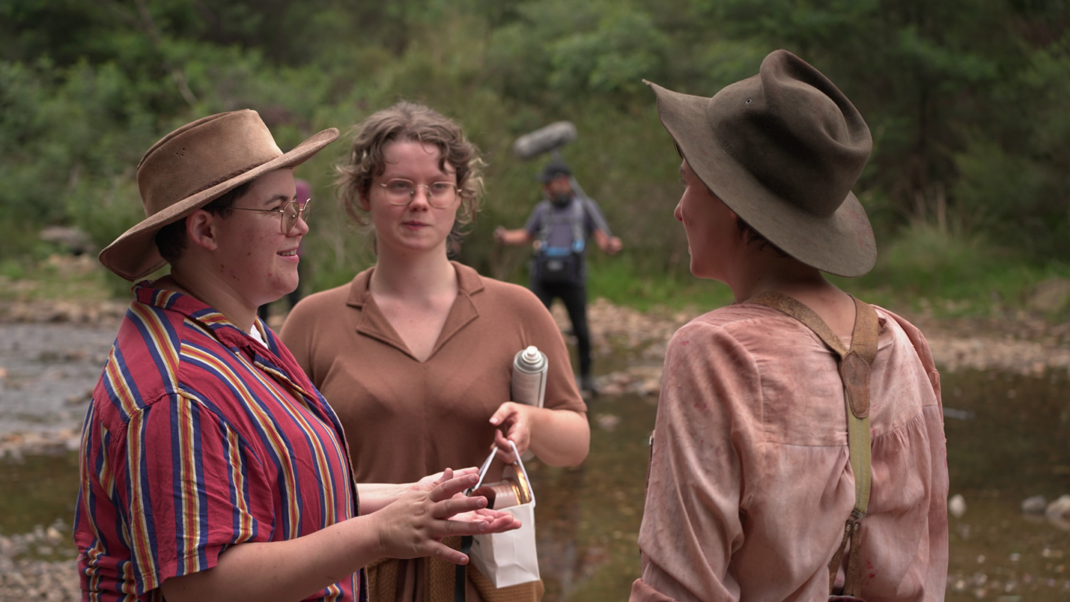 Three people wearing hats speak in front of a river, in the background a man holds a boom mic.