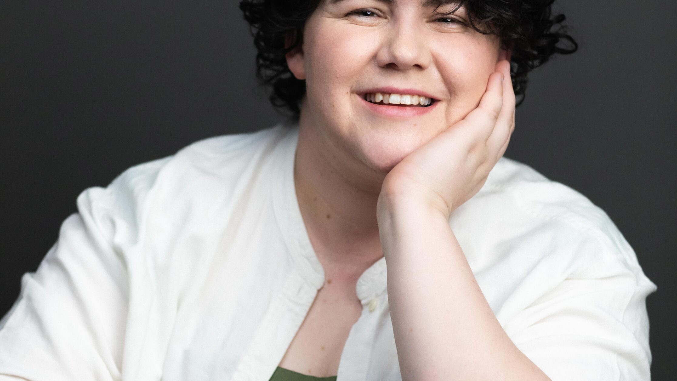 Person with curly black hair leans their chin on one hand, their arm resting on a table. they are wearing a white shirt with a green singlet underneath and smiling.