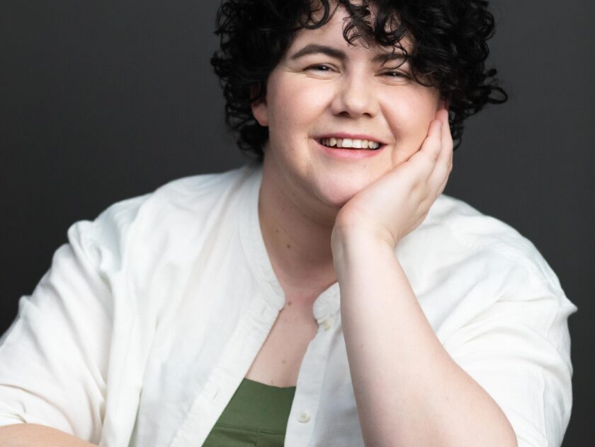 Person with curly black hair leans their chin on one hand, their arm resting on a table. they are wearing a white shirt with a green singlet underneath and smiling.