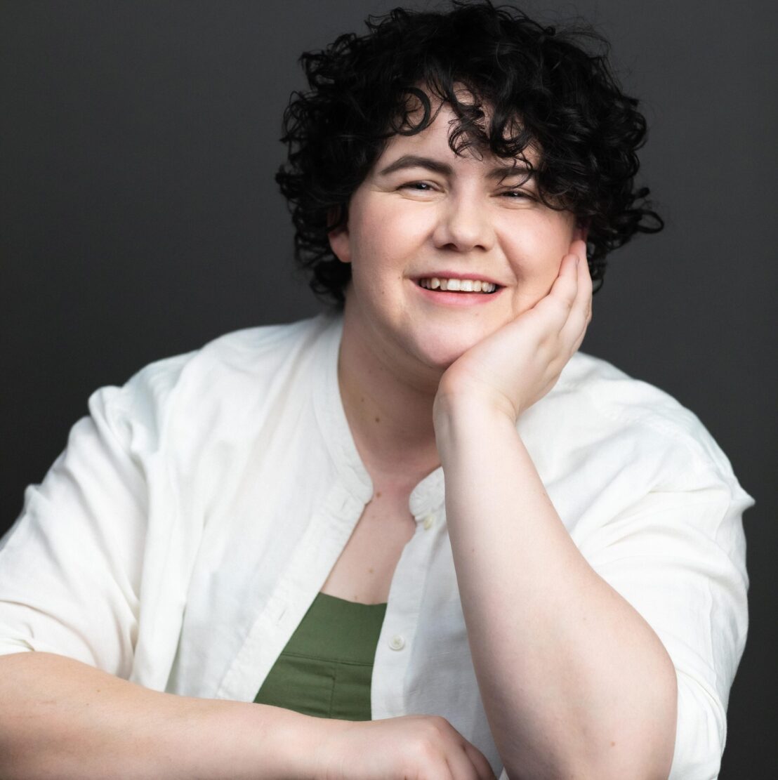 Person with curly black hair leans their chin on one hand, their arm resting on a table. they are wearing a white shirt with a green singlet underneath and smiling.