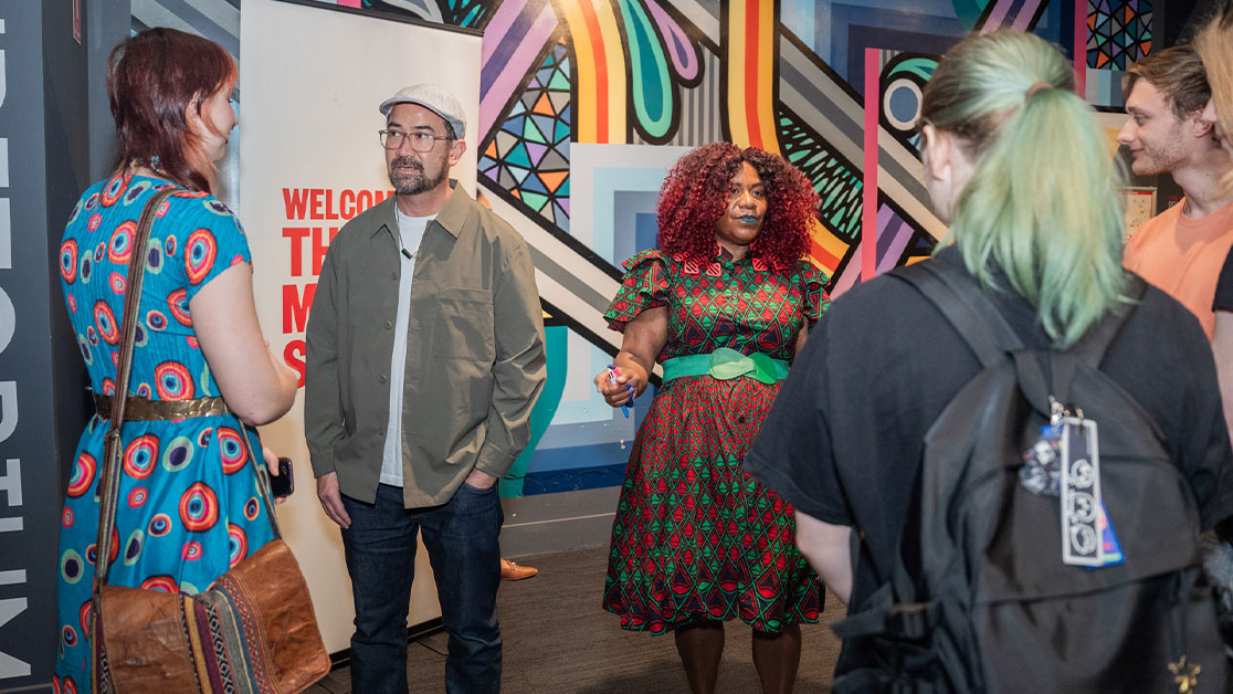 man and woman speak to group of young people in front of colourful backdrop wall