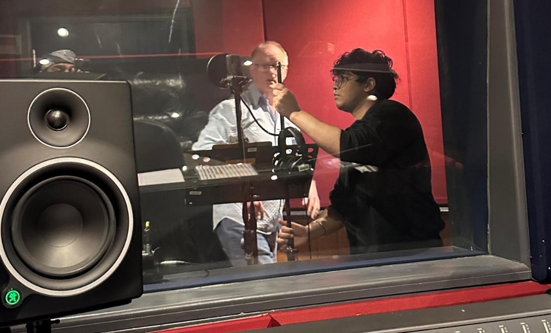 person with curly hair and glasses helps middle aged man with microphone in recording booth