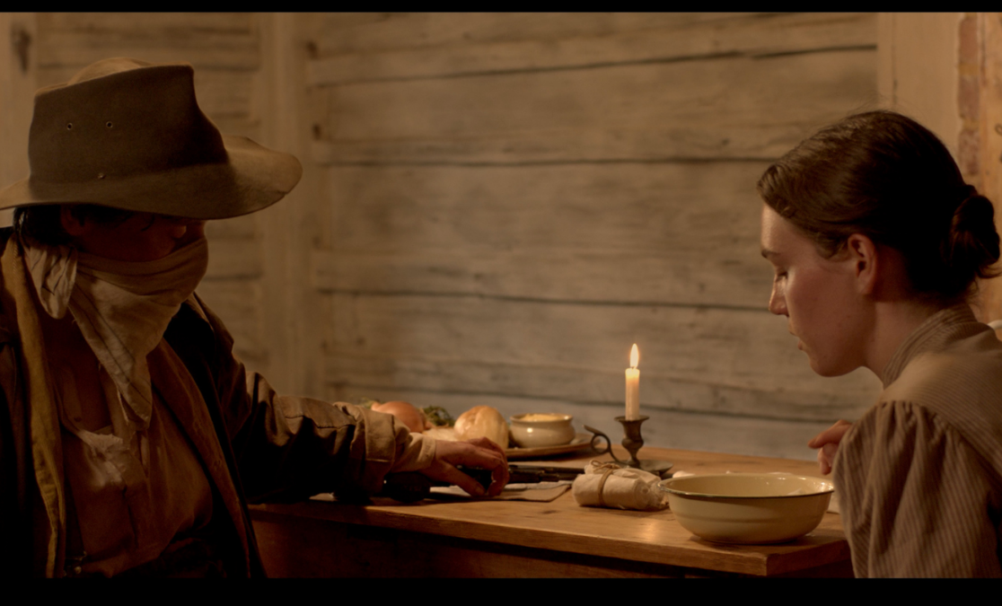 A person dressed in bushranger clothes with their face covered faces a woman in a colonial australian kitchen with a candle in the background