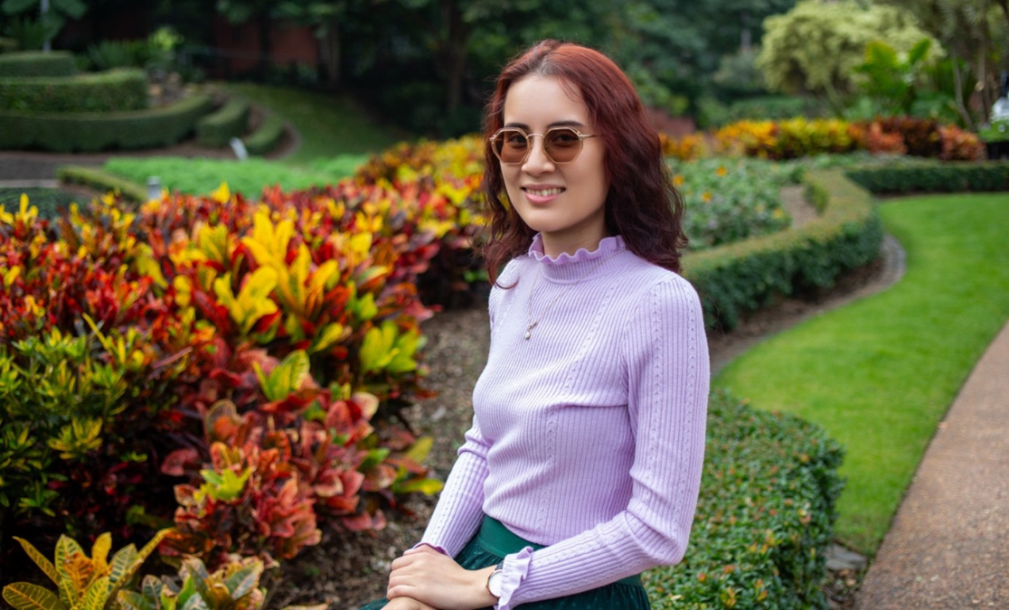 Young woman with red hair wearing glasses in purple top stand side on in front of colourful flower garden