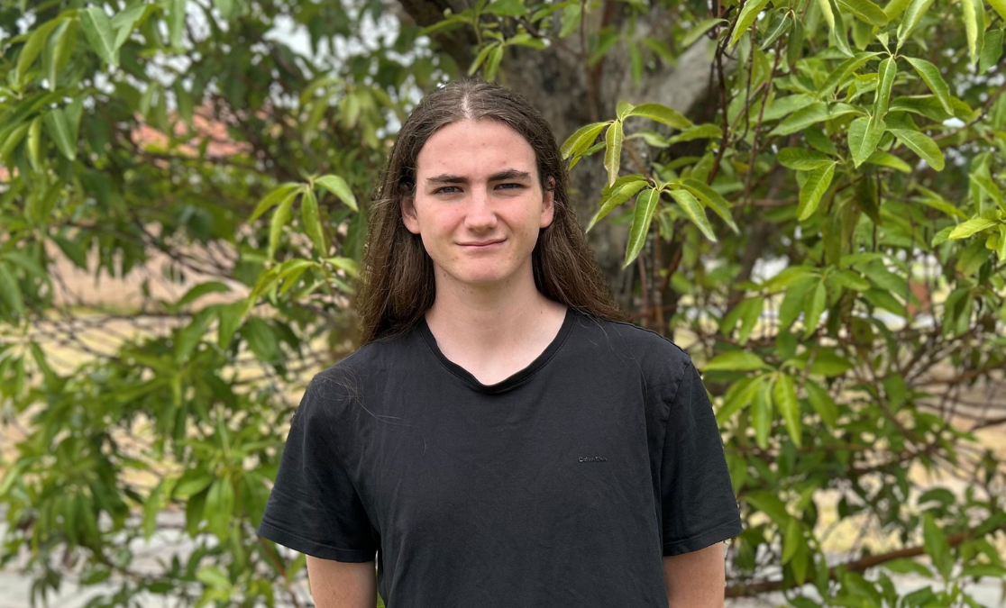 young man with logn hair stand in front of a bush in a black t shirt smiling