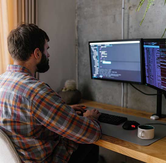 Man with beard wearing a flannel shirt sits at desk with dual monitors. Code on screen. He has both hands on keyboard and looking at screen.