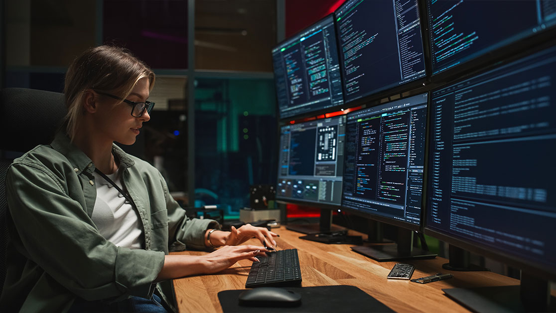 A person with long hair and glasses sits at desk looking at keyboard. 6 monitors are placed in front of the person at the desk.