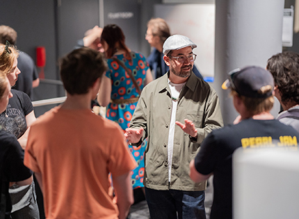 An SAE University College teacher wearing classes and a flat cap showing students around a college event - tile image 430x316