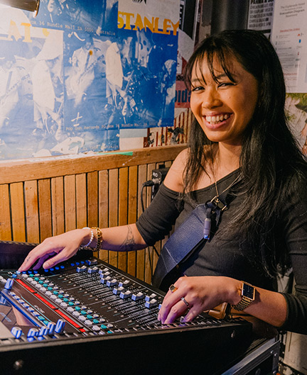 SAE Bachelor Audio Engineering alumni Aiesha Bacalso using a mixing desk in a room with wooden panelling and smiling towards camera - mobile image