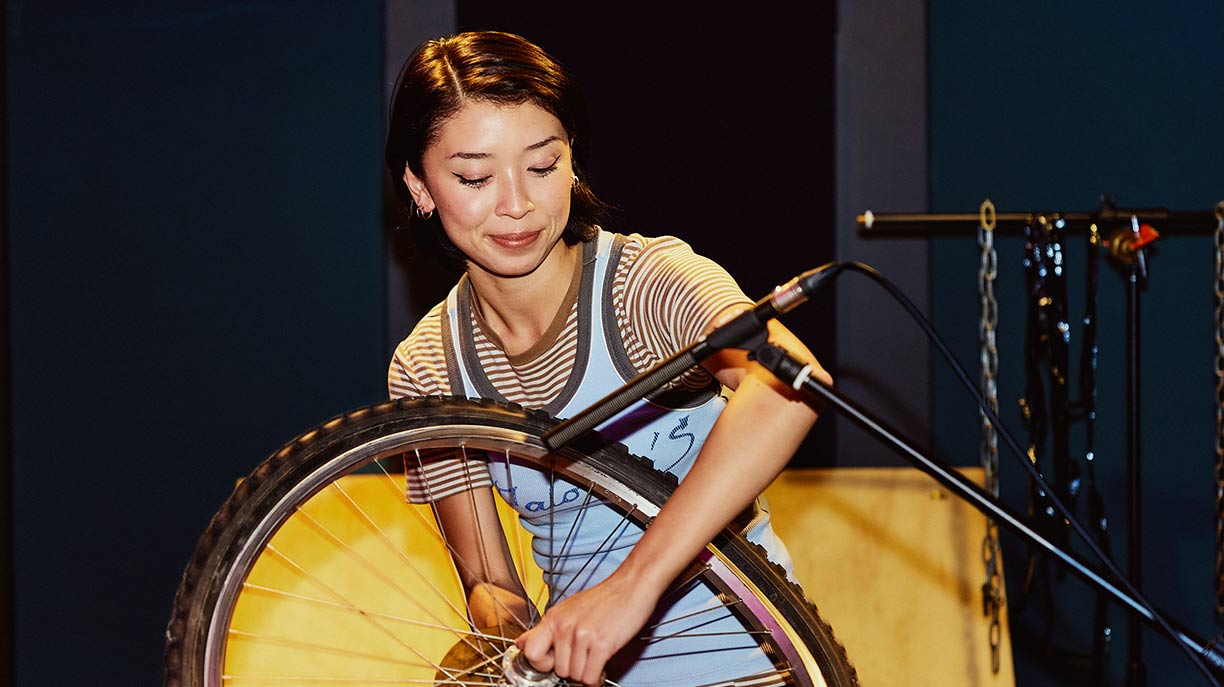 A young female student standing in a dark recording studio, holding a bicycle wheel up to a microphone