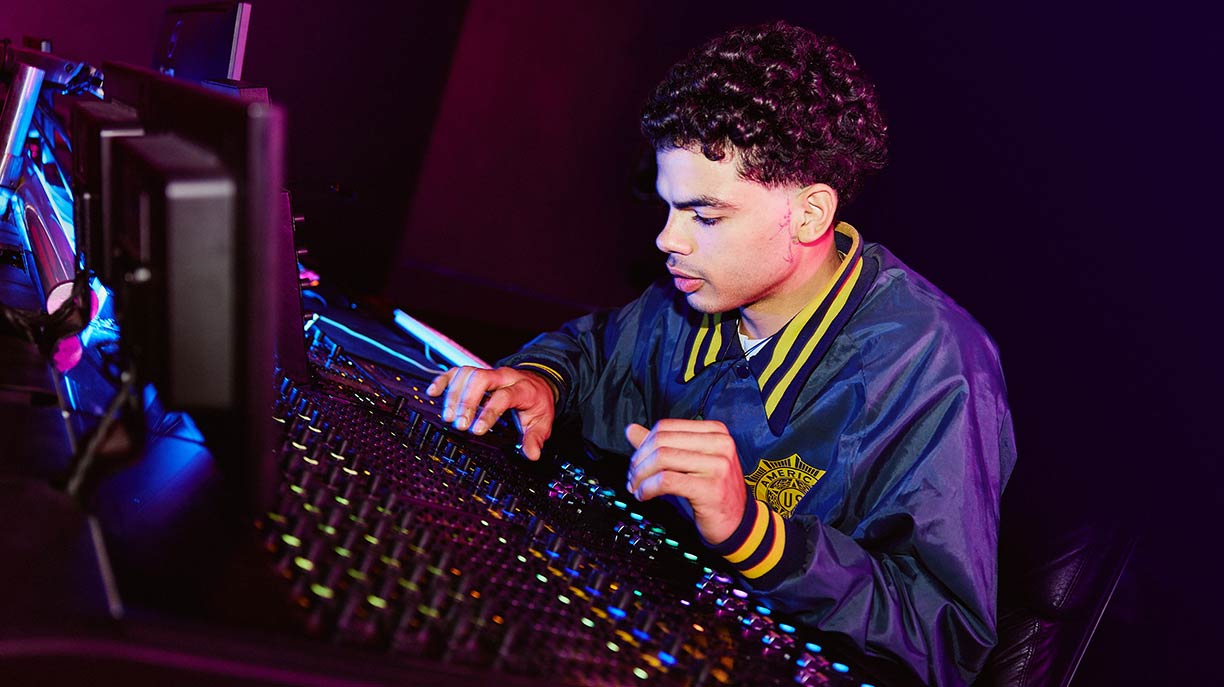 A young male student operating a mixing desk in a room with dark lighting 1224x687