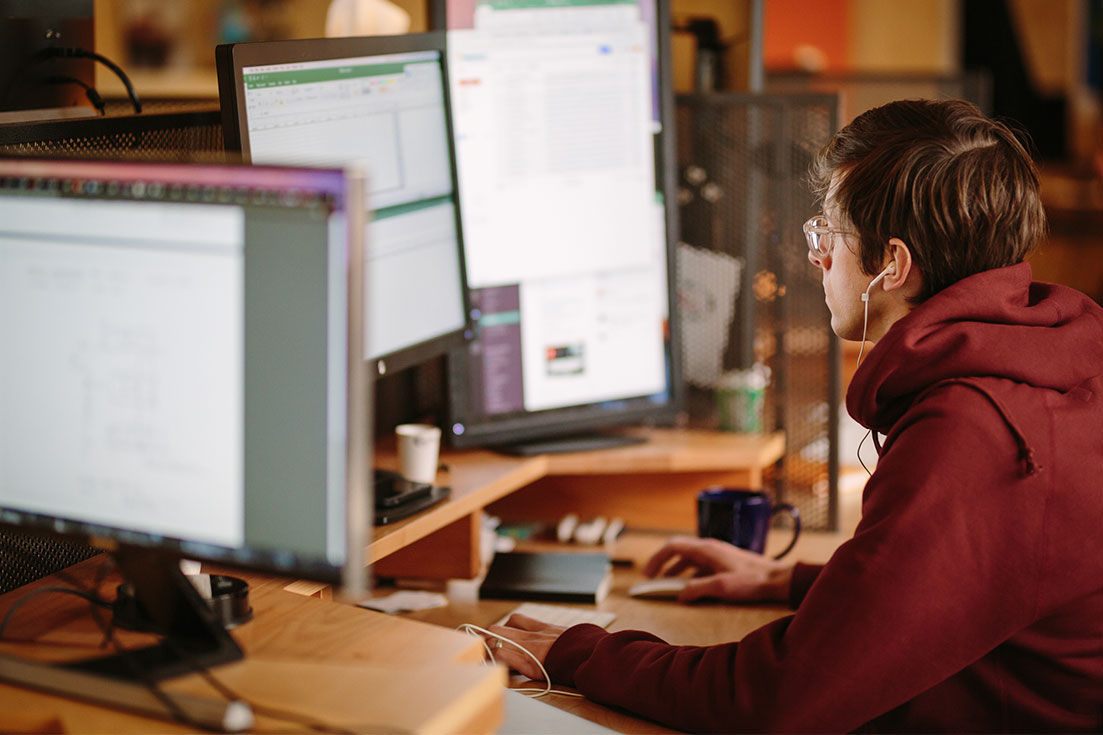 Guy wearing wired headphones sits at monitor and desk. He is at his desk, eyes on screen, hand on mouse and keyboard.
