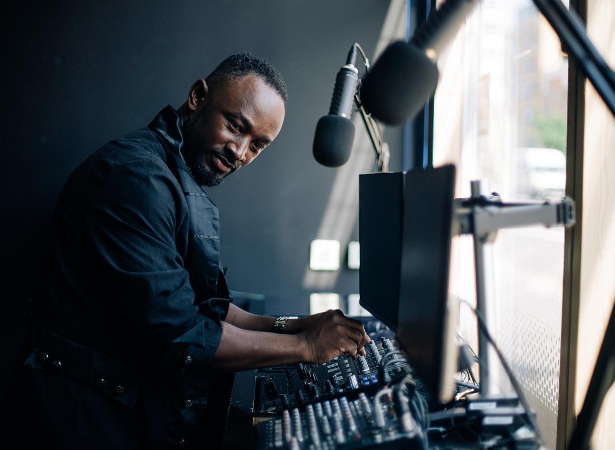 A man operating a channel mixer in front of two microphones, two monitors and other audio hardware