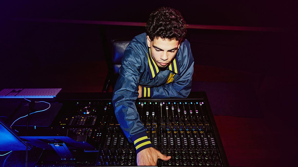 A young male student sitting at a mixing desk in a dark room, reaching forward towards the desk with is right hand.