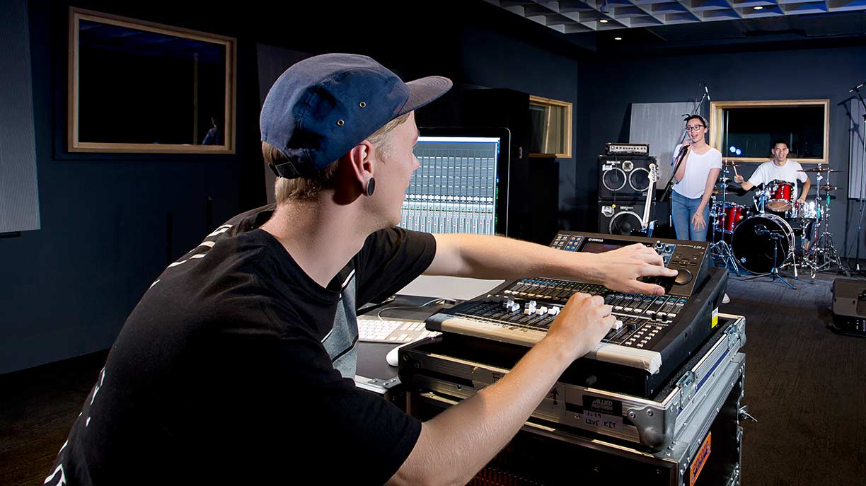 A male student wearing a hat using audio production equipment while a band plays live music in the background