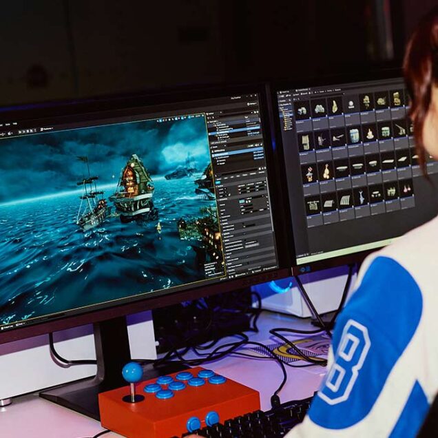 A woman with red hair in a ponytail sitting at a desk facing away from camera, working with games software on a desktop monitor