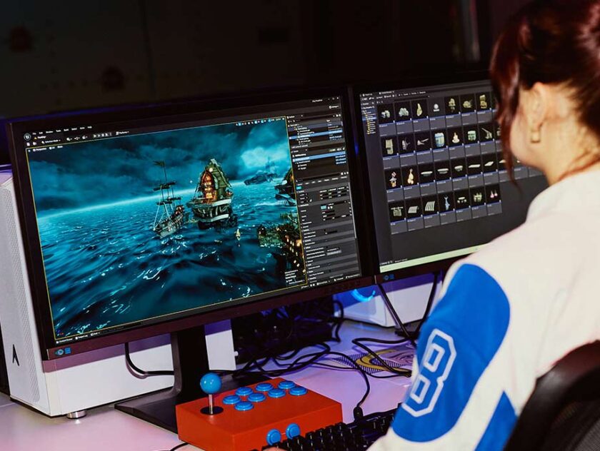 A woman with red hair in a ponytail sitting at a desk facing away from camera, working with games software on a desktop monitor