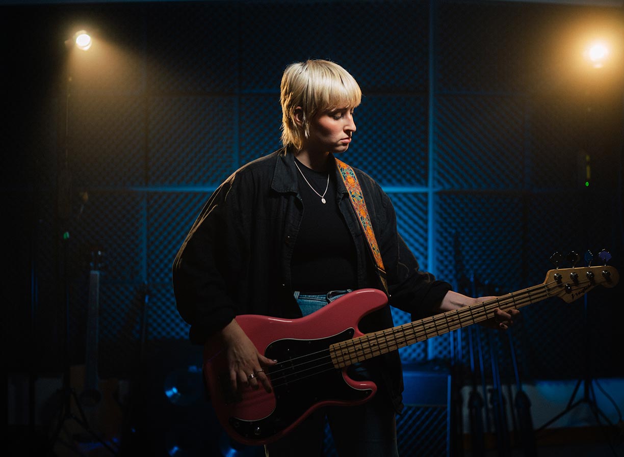 A student with short blonde hair playing a bass guitar in a studio with blue lighting