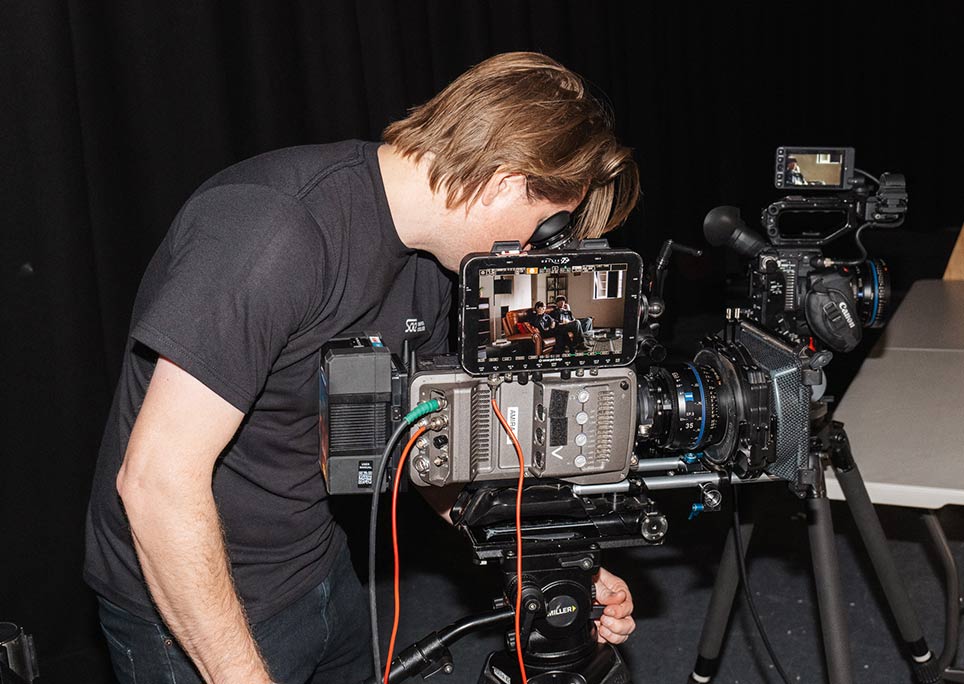 A man in all black clothing leaning downwards to operate a film camera, standing side-on to camera
