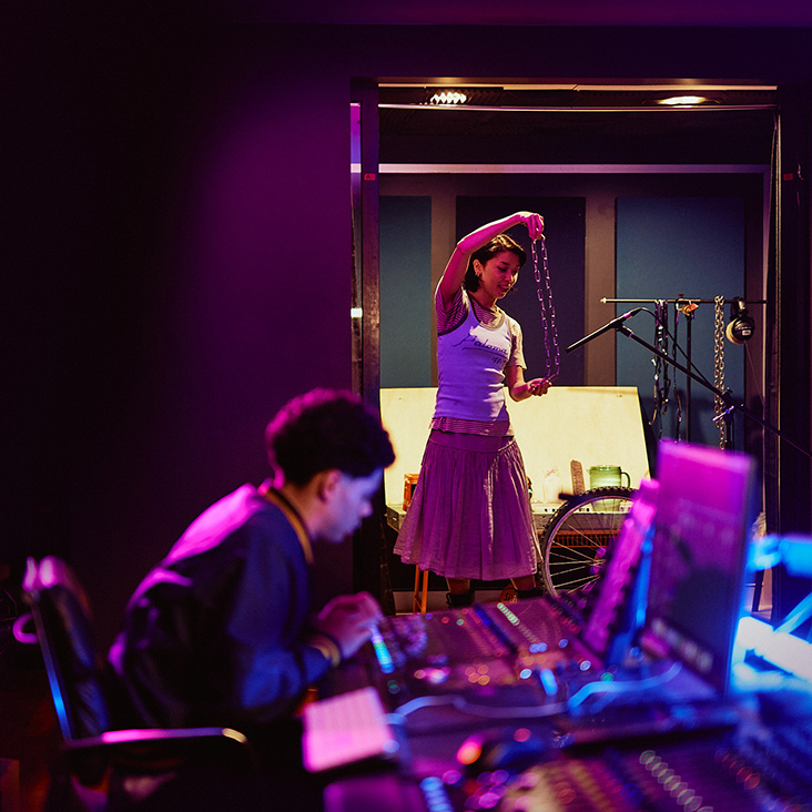 A male student sitting at a desk with audio hardware and female student in the background holding a chain up to a microphone