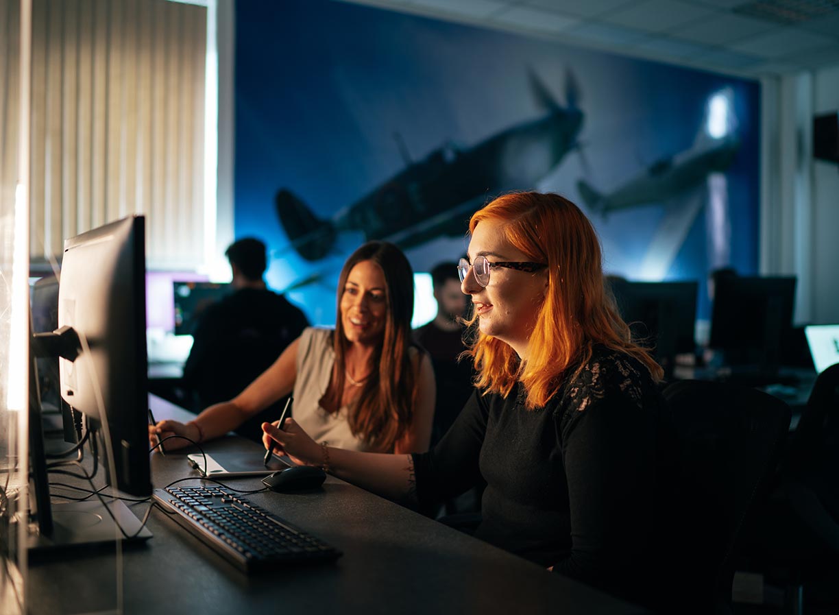 Two female animation students facing side on to camera, sitting at a desk and looking towards a desktop computer monitor