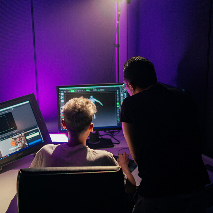 Two students at a desk with their backs faced towards camera, working on an animation project on two monitors in a room with purple backlighting