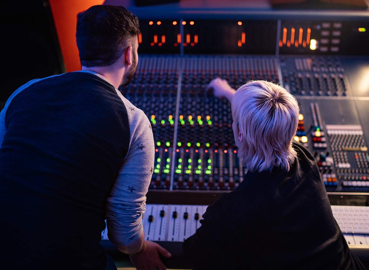 Two SAE Audio students sitting at a mixing desk facing with their back to camera, with one pointing towards the desk