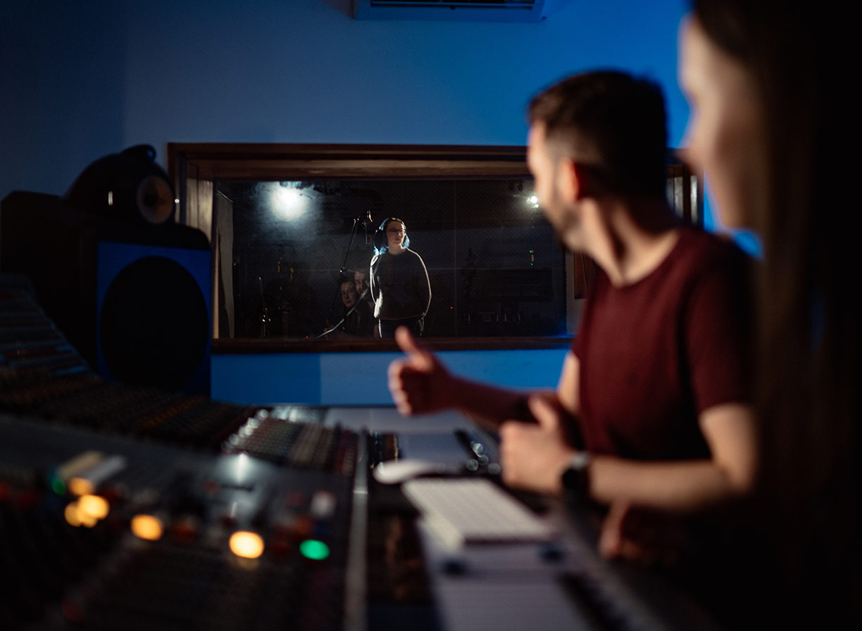 Two SAE Audio students sitting in front of audio hardware with a female student standing in a recording booth in the background