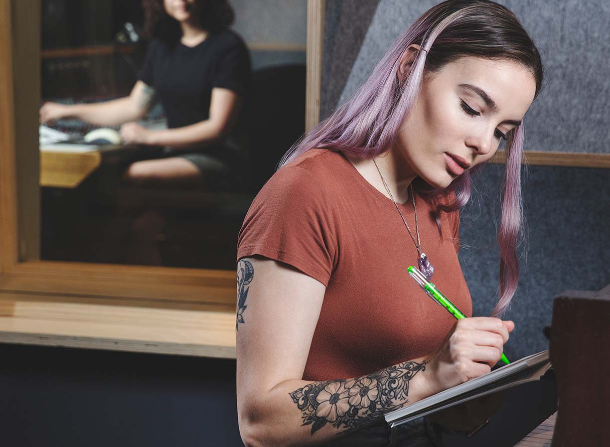 A female student writing in a notepad in front of a piano, with an older female teacher watching on