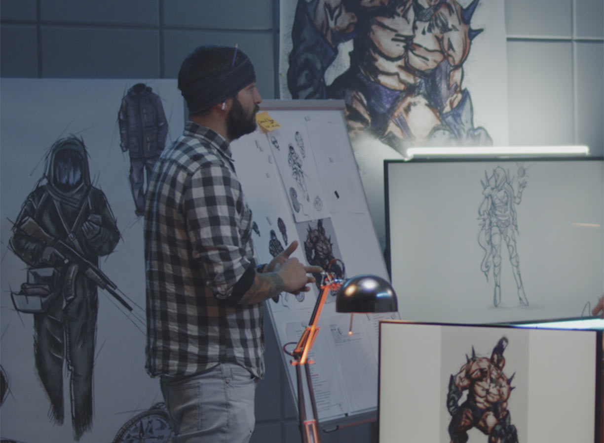 A male student wearing a black beanie and standing in front of an easel and monitor with illustrations on them