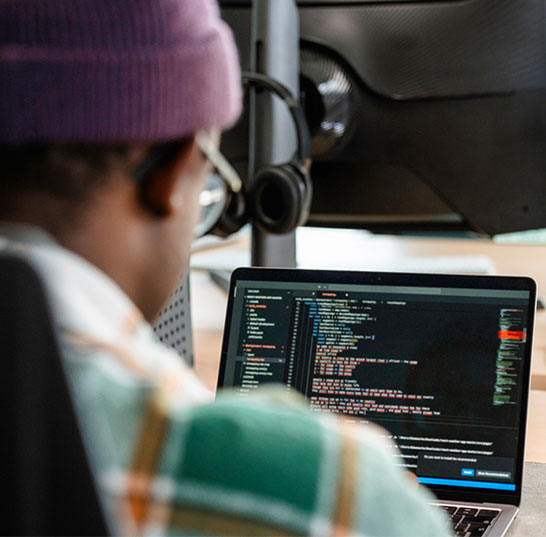 Person wearing a beanie, flannel shirt and glasses sits in front of laptop full of code.