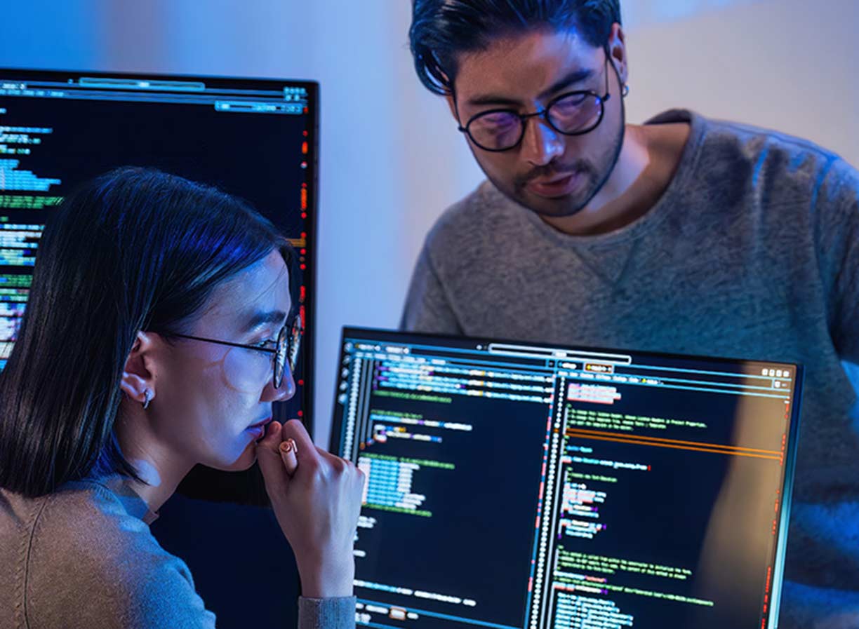 A female analyst programmer sitting at a desk in front of two computer monitors, with a male programmer watching on - Information Technology