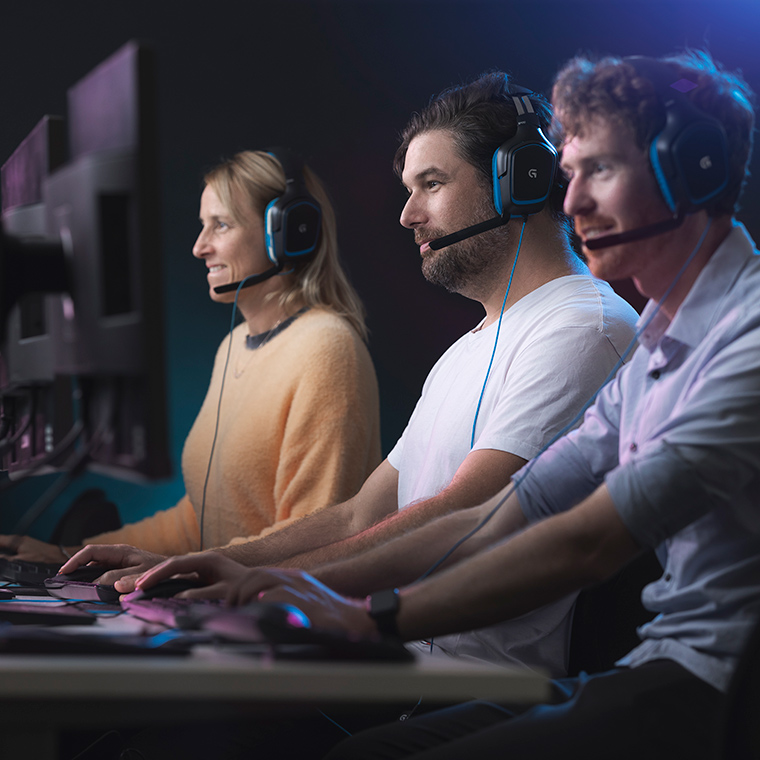Three games development students sitting at computers and wearing headset, facing side on to camera