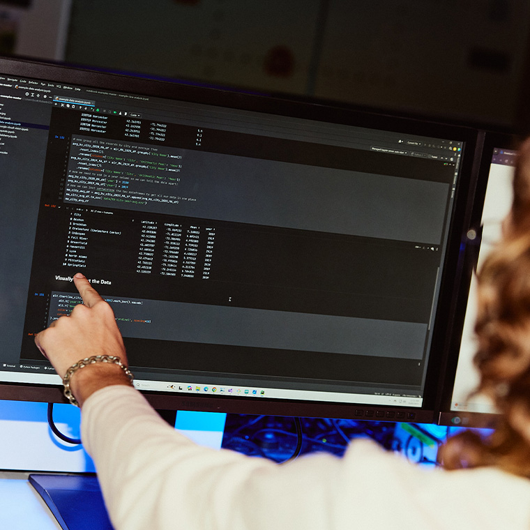 A woman's hand pointing towards a desktop monitor with software displaying a code editor - Information Technology