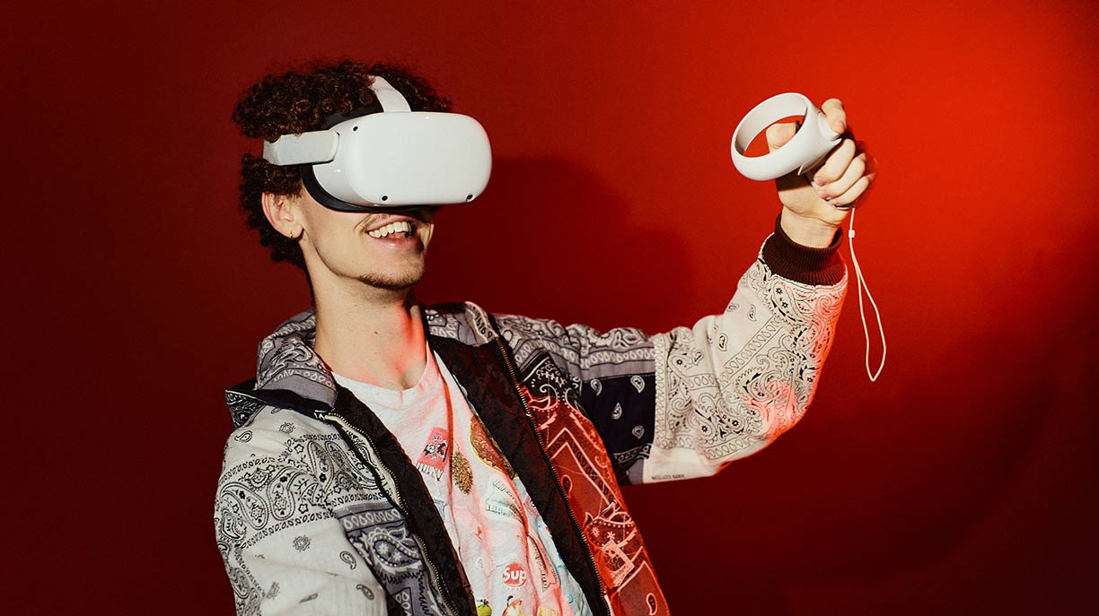 A smiling man wearing a virtual reality headset and holding holding a controller in the air with his left hand in front of a red background