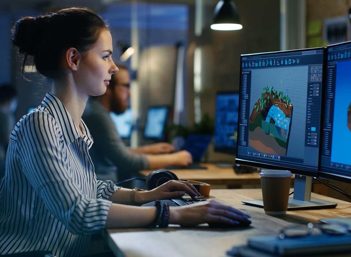 A female 3D modeller sitting at a desk working on two monitors displaying games development software, sitting side on to camera
