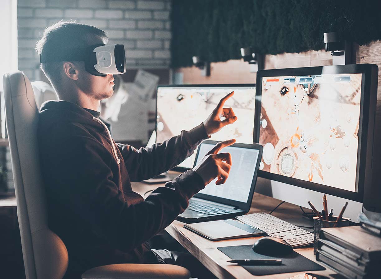 A male game designer sitting at a desk in front of several monitors, wearing a virtual reality headset and holding both arms in the air with index fingers pointed