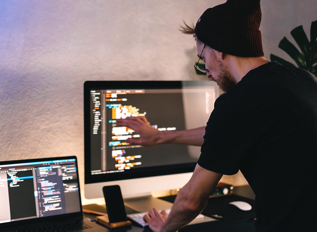 A male game programmer wearing a black beanie and glasses, standing over a desk with one hand on a computer keyboard and one hand pointing towards a monitor displaying development code
