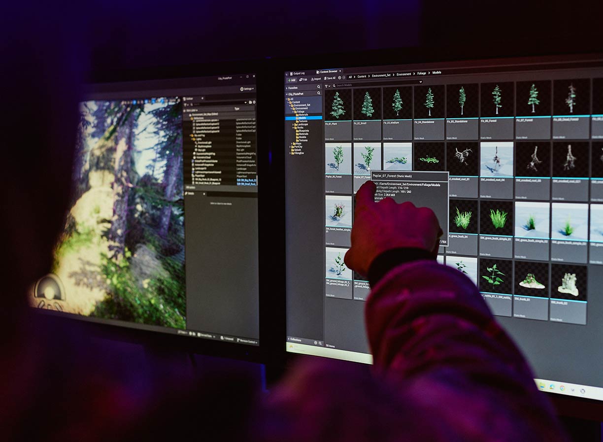 The hand of of a male level designer pointing towards two monitors displaying various graphics of plant life