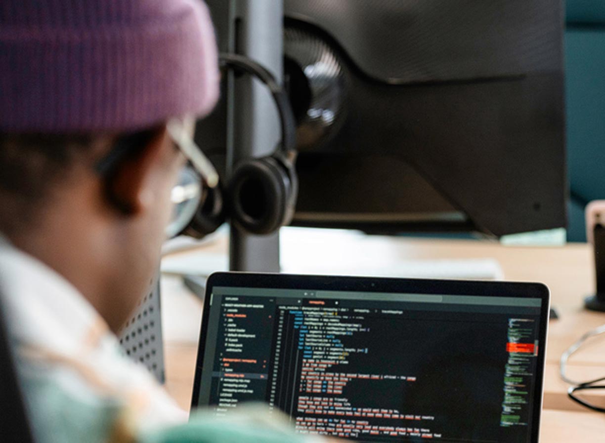 A man wearing a purple beanie and glasses working on software programming on a laptop computer - Information Technology