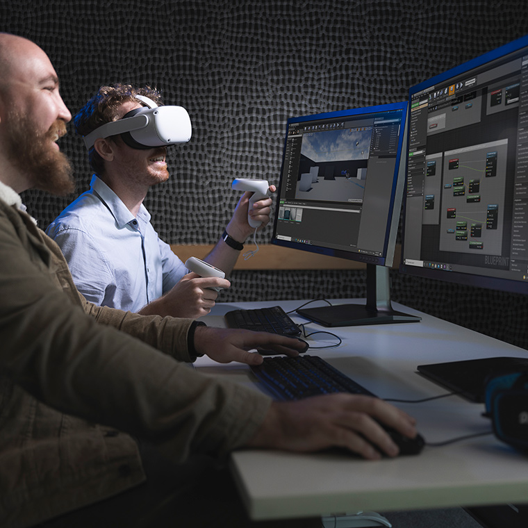 Two male games development students sitting in front of two large monitors. One student is operating the mouse while the other student wears a virtual reality headset and holds controllers in either hand.