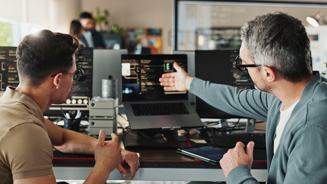 A male teacher and student sitting in front of various computer hardware, with the teacher gesturing towards a laptop screen with an open hand