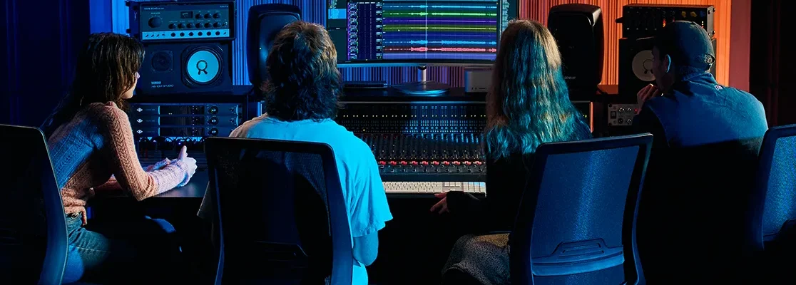 Four students gather in front of a console and editing software on monitor. SAE is now a University College