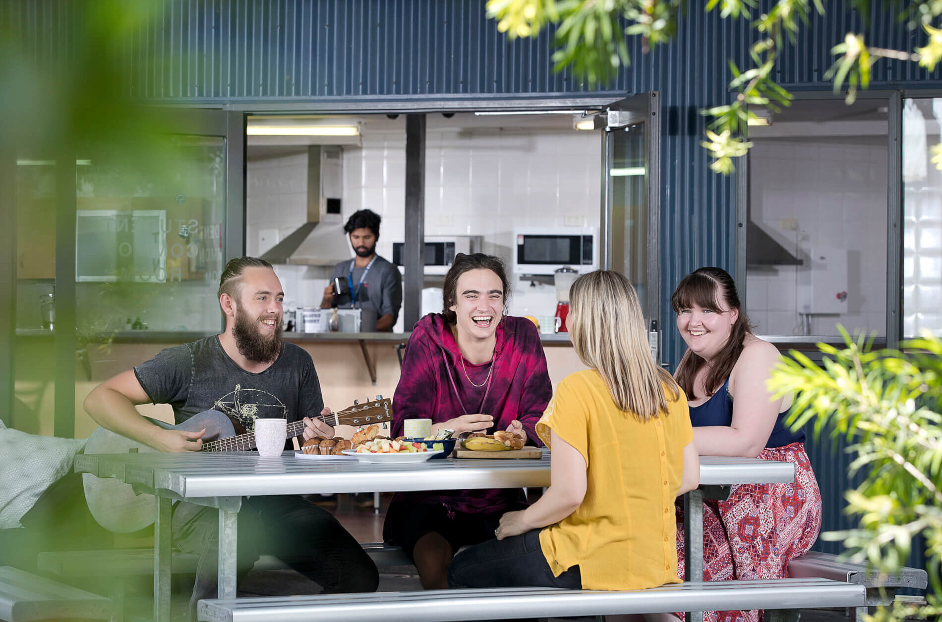 A group of four people sit at a table in outside Common Area. Food and coffee cups are on the table. One person plays an acoustic guitar