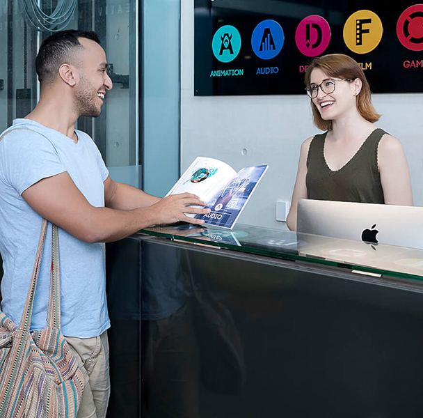 A man wearing a bag and holding an open book, standing at a reception desk and being assisted by a student advisor