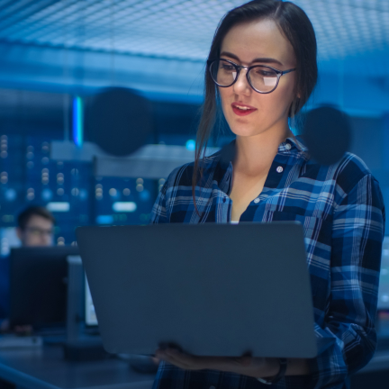 A woman with glasses standing, working on a laptop in room with blue lighting
