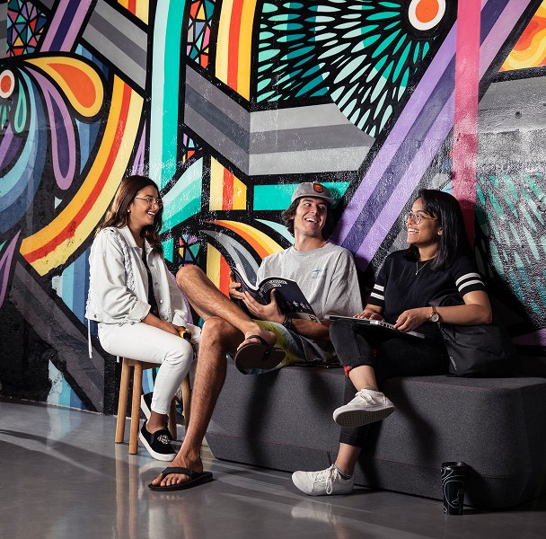 Three students talking on a lounge, sitting in front of a colourful mural